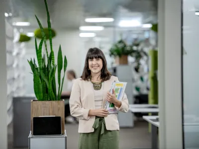 Selina Infanger steht lächelnd in einem modernen Büro. Sie trägt eine helle Jacke, ein beigefarbenes Oberteil und weite grüne Hosen. In ihren Armen hält sie mehrere Dokumente und Unterlagen. Im Hintergrund befinden sich Büropflanzen, darunter eine hohe Grünpflanze in einem Holztopf, der auf einem kleinen Regal steht, sowie weitere Dekorationselemente und Möbel. Die Büroeinrichtung wirkt hell und offen, mit modernen Gestaltungselementen und einer klaren, strukturierten Umgebung. 