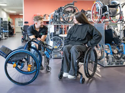 Chiara sitzt vor einem Regal voller Rollstühle in ihrem Rollstuhl, neben ihr kniet ein Mitarbeiter der Orthotec am Boden und hält einen Rollstuhl gekippt, um Chiara daran etwas zu zeigen.