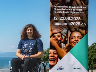 Giulia Damiano sitzt in ihrem Rollstuhl vor dem Genfersee und trägt ein T-Shirt mit der Aufschrift "Lausanne 2025". Rechts von ihr ist die Werbung für das Eidgenössische Turnfest in Lausanne. 