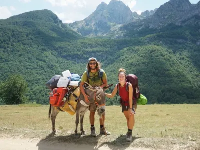 Eine Frau und ein Mann stehen mit einem bepackten Esel in einer grünen Berglandschaft.