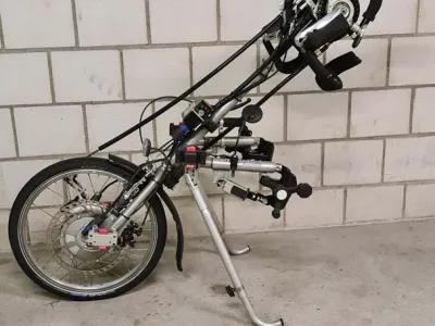 Rollstuhlvorsatz mit Handkurbel an weißer Wand lehnend.