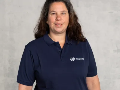Ein freundliches Porträt einer Frau mit langen braunen Haaren, die einen dunkelblauen Poloshirt mit einem weissen Logo trägt.