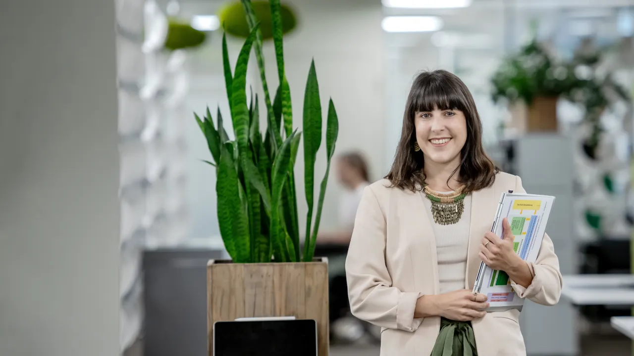 Selina Infanger steht lächelnd in einem modernen Büro. Sie trägt eine helle Jacke, ein beigefarbenes Oberteil und weite grüne Hosen. In ihren Armen hält sie mehrere Dokumente und Unterlagen. Im Hintergrund befinden sich Büropflanzen, darunter eine hohe Grünpflanze in einem Holztopf, der auf einem kleinen Regal steht, sowie weitere Dekorationselemente und Möbel. Die Büroeinrichtung wirkt hell und offen, mit modernen Gestaltungselementen und einer klaren, strukturierten Umgebung. 
