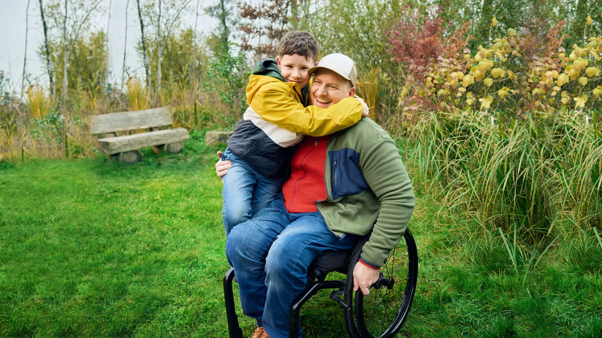 Heiri sitzt in seinem Rollstuhl auf einer Wiese und sein Sohn klettert ihm halbwegs auf den Schoss und umarmt seinen Vater ganz fest. Beide lächeln.