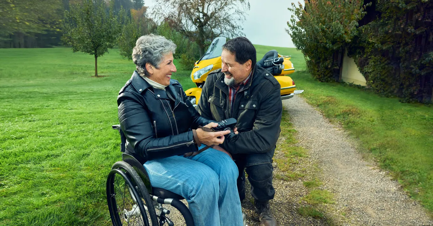 Rosa Zaugg sitzt im Rollstuhl auf einem Kiesweg, hält eine Kamera in der Hand und lächelt ihren Partner Lukas an.