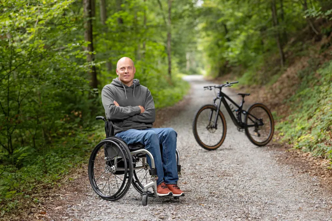 Sebastian Tobler est en fauteuil roulant dans la forêt et sourit dans la caméra, les bras croisés. À l’arrière-plan se trouve un VTT. 
