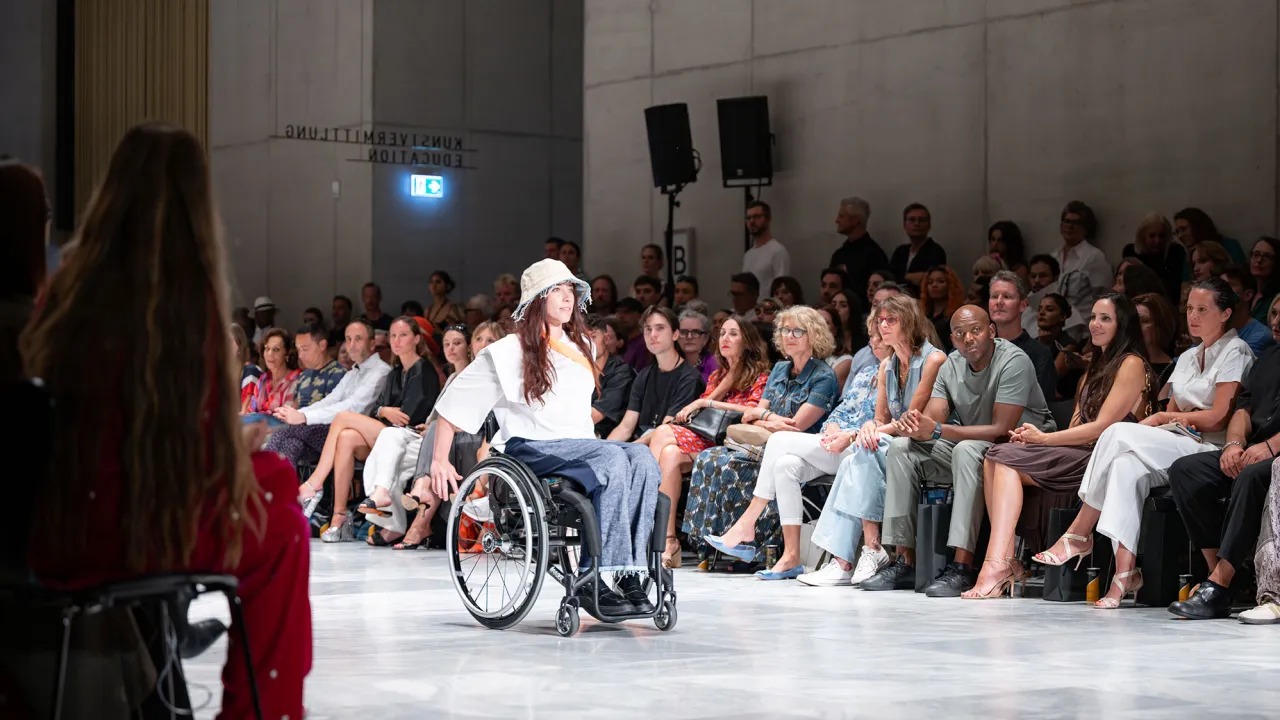 Modeshow mit Rollstuhlfahrerin auf dem Laufsteg die inklusive Mode trägt.