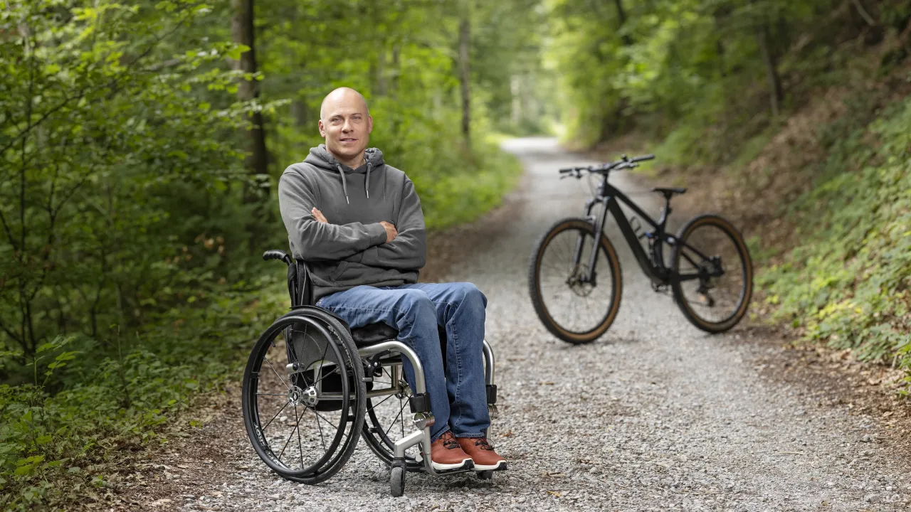 Sebastian Tobler sitzt in seinem Rollstuhl im Wald und lächelt mit verschränkten Armen in die Kamera. Im Hintergrund ist ein Mountain Bike zu sehen. 