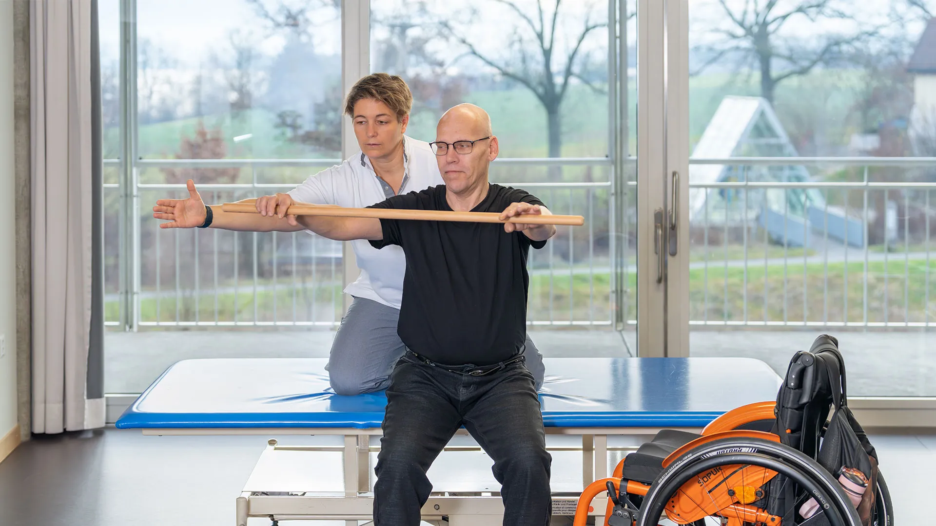 Ein Mann sitzt auf einem Liegebett mit seiner Therapeutin hinter ihm und macht eine Übung.