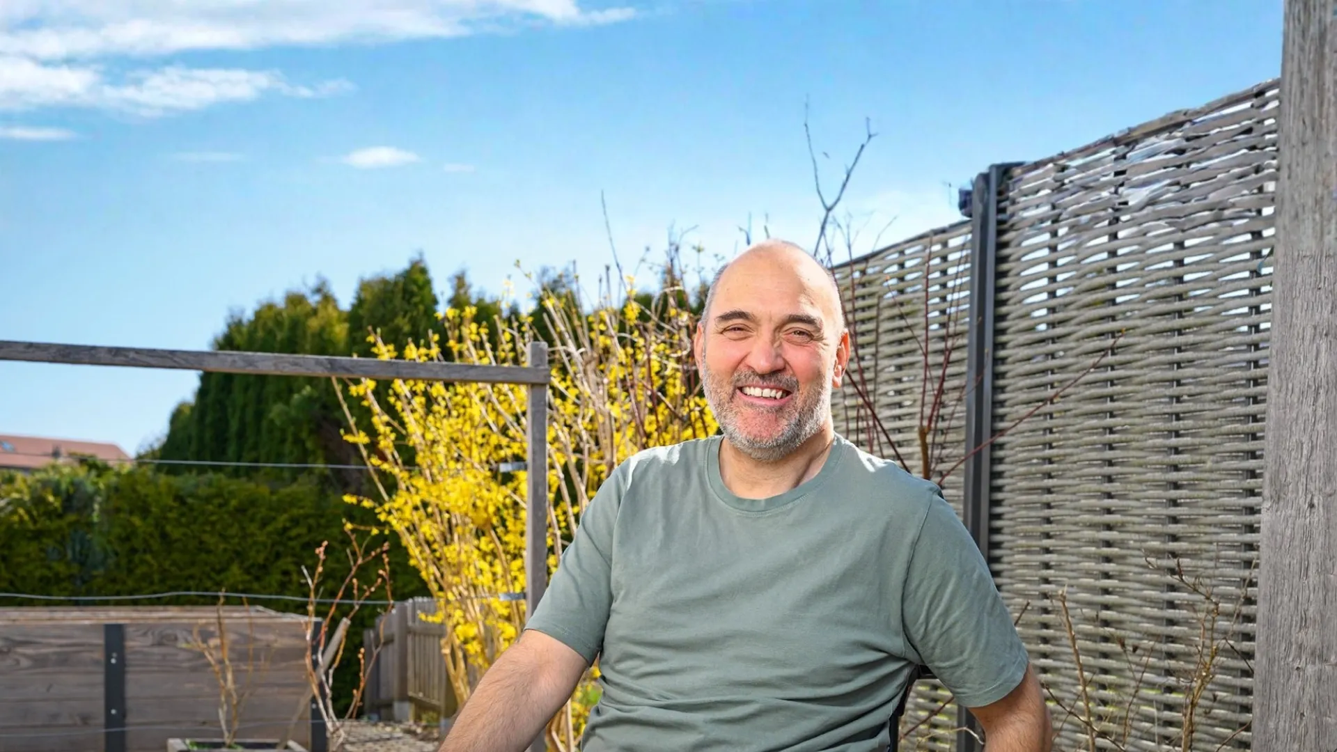Lächelnder Mann im Rollstuhl sitzt bei sonnigem Wetter auf einer Terrasse im Garten – selbstbestimmtes Leben, Barrierefreiheit und Lebensfreude im Alltag.