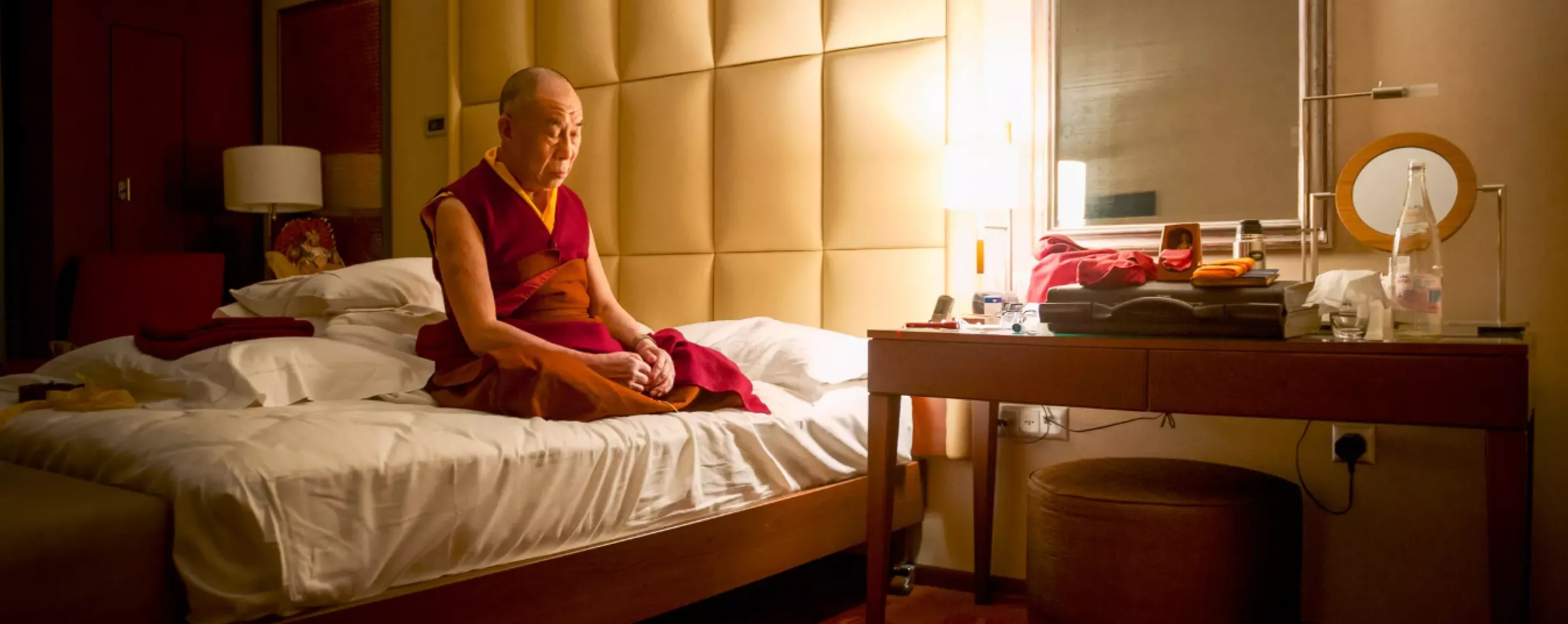Dalai Lama sitting on bed, reading book in warm, cozy bedroom.