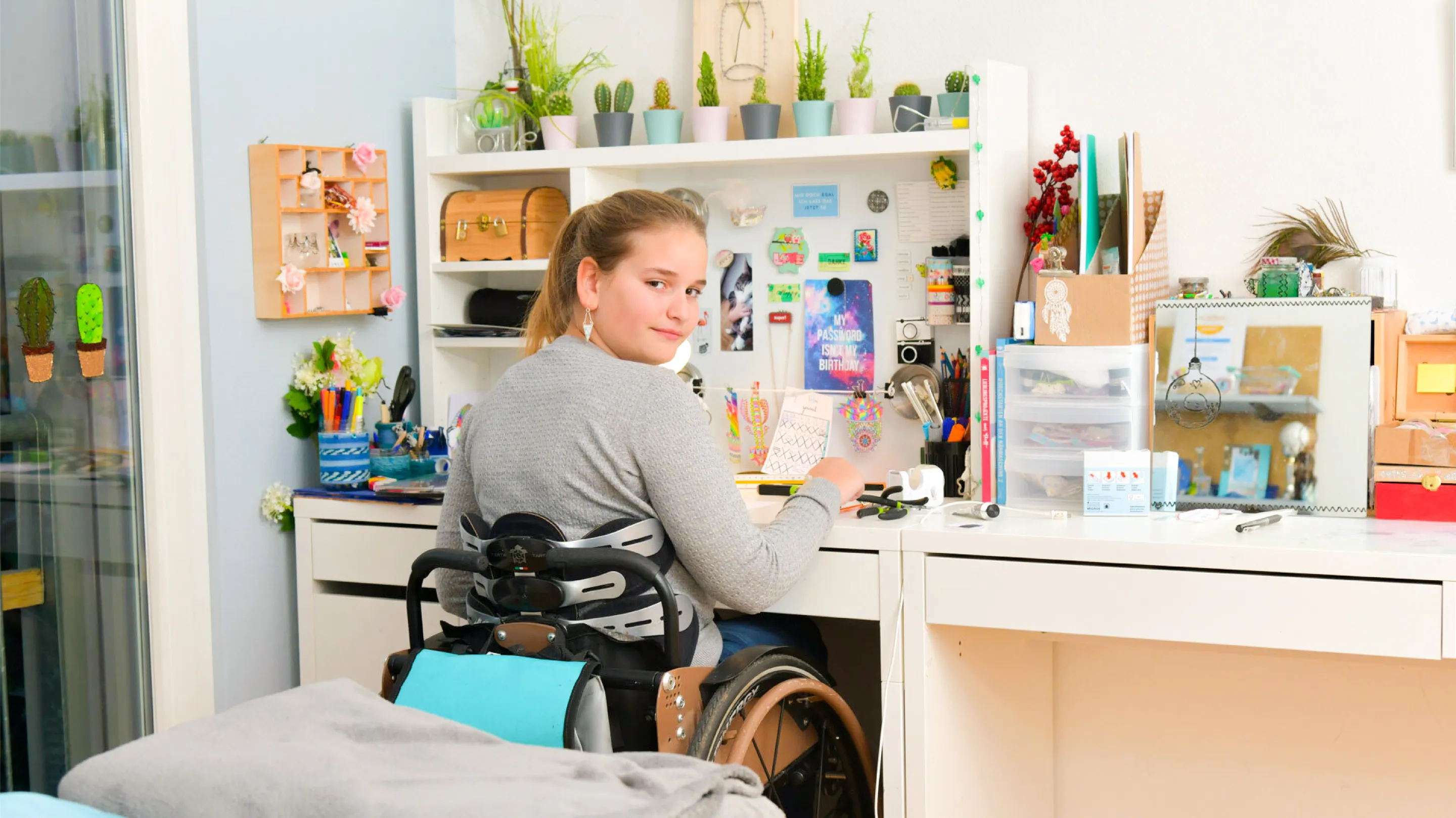 A girl is sitting in a wheelchair at her desk and looking over her shoulder into the camera. 