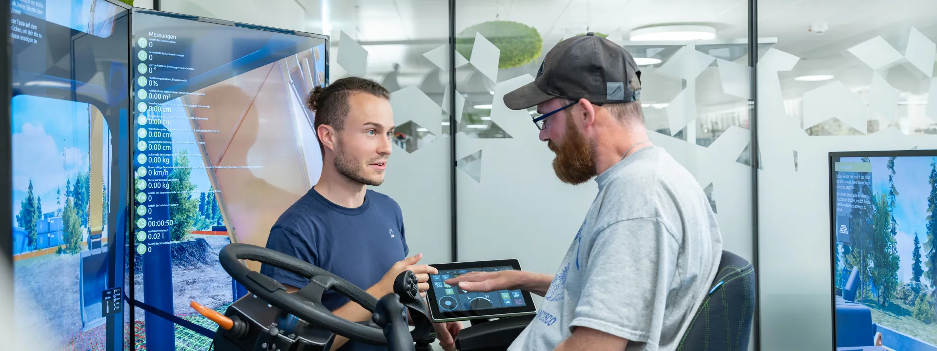 Ein Mann im Rollstuhl steuert einen Baumaschinensimulator und ein Mann erklärt es ihm.