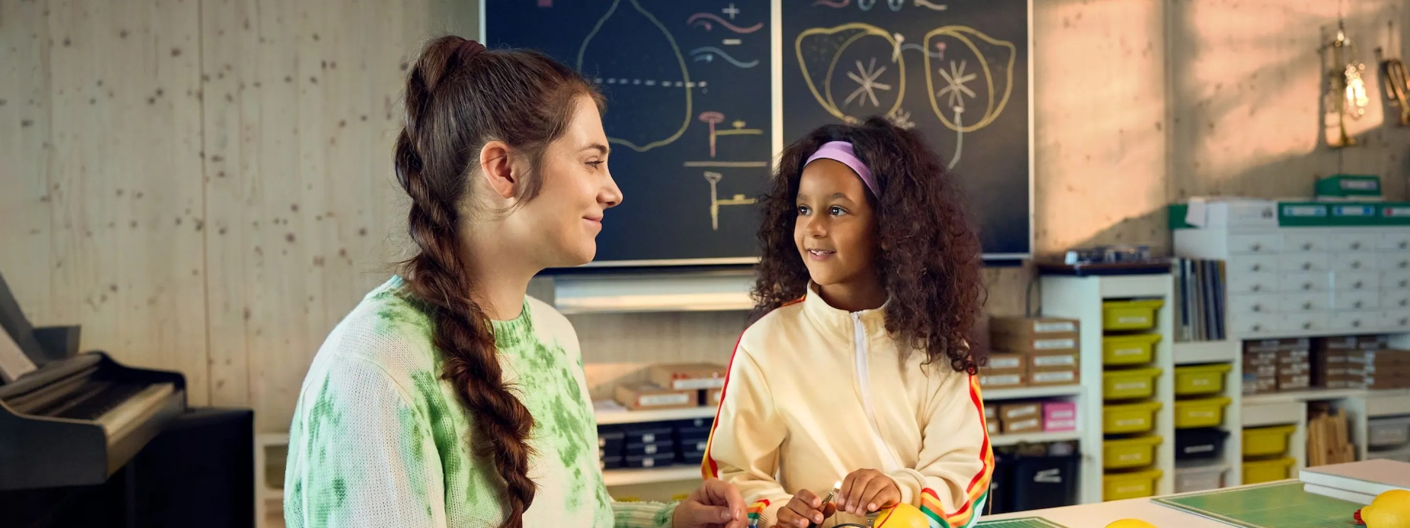 Chiara Schlatter sitzt im Rollstuhl an einem Tisch in einem Schulzimmer zusammen mit einer Schülerin und zeigt ihr, wie man mit Zitronen eine Lampe zum Leuchten bringen kann. Beide schauen sich an und lächeln.