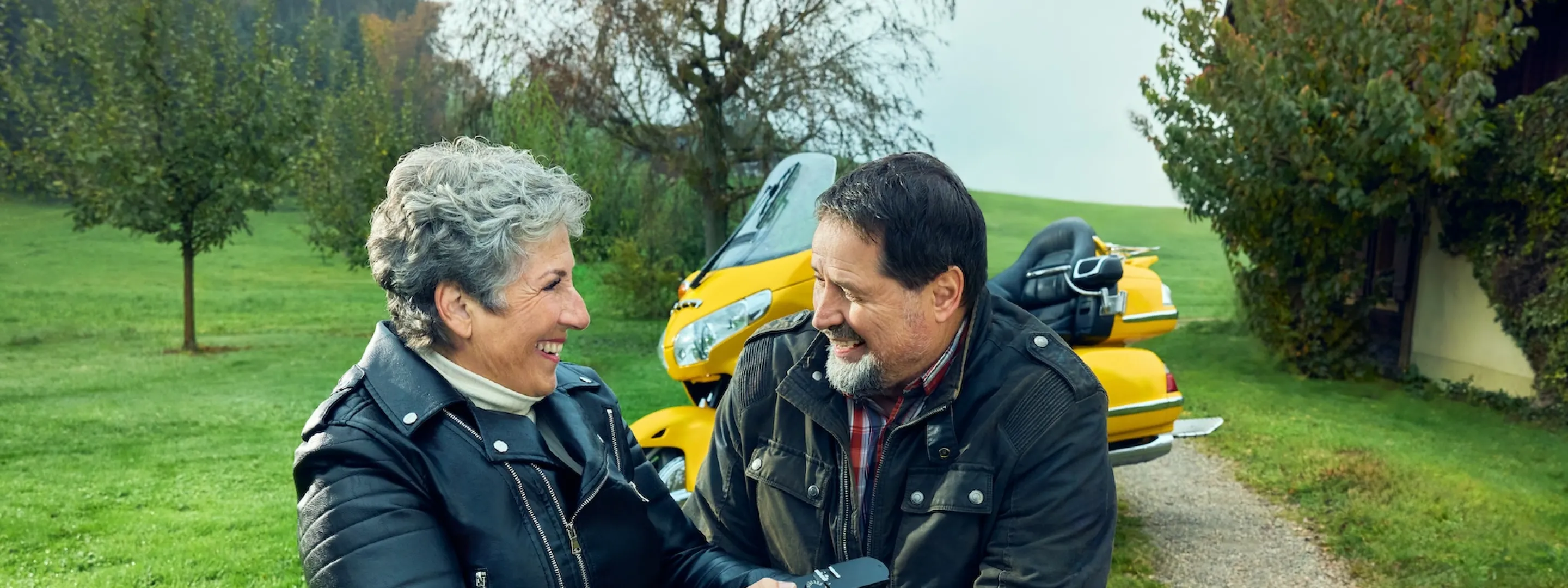 Rosa Zaugg sitzt in ihrem Rollstuhl auf einem Feldweg auf einem Bauernhof und blickt zu ihrem Partner Lukas hinüber, der sich neben ihr hingekniet hat. Sie zeigt ihm eine Kamera. Im Hintergrund ist seine gelbe Goldwing zu sehen.