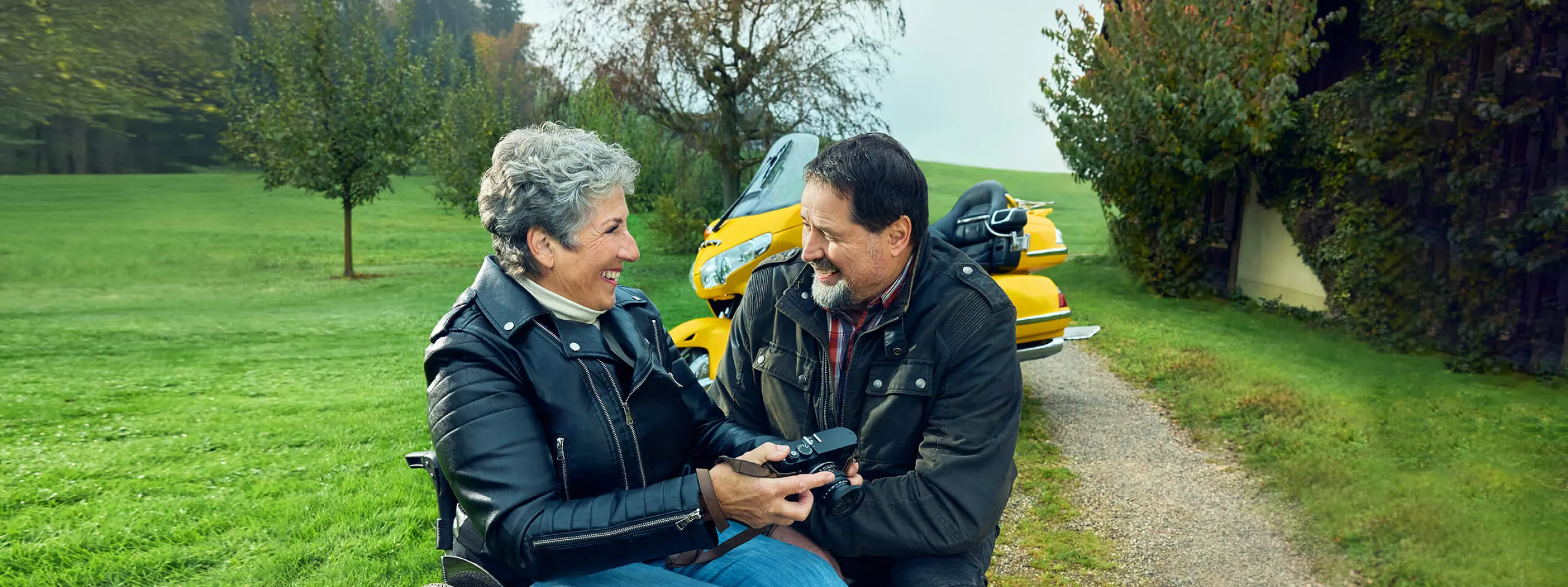 Rosa Zaugg è seduta in carrozzina su un vialetto di ghiaia, tiene in mano una macchina fotografica e sorride al suo partner Lukas.