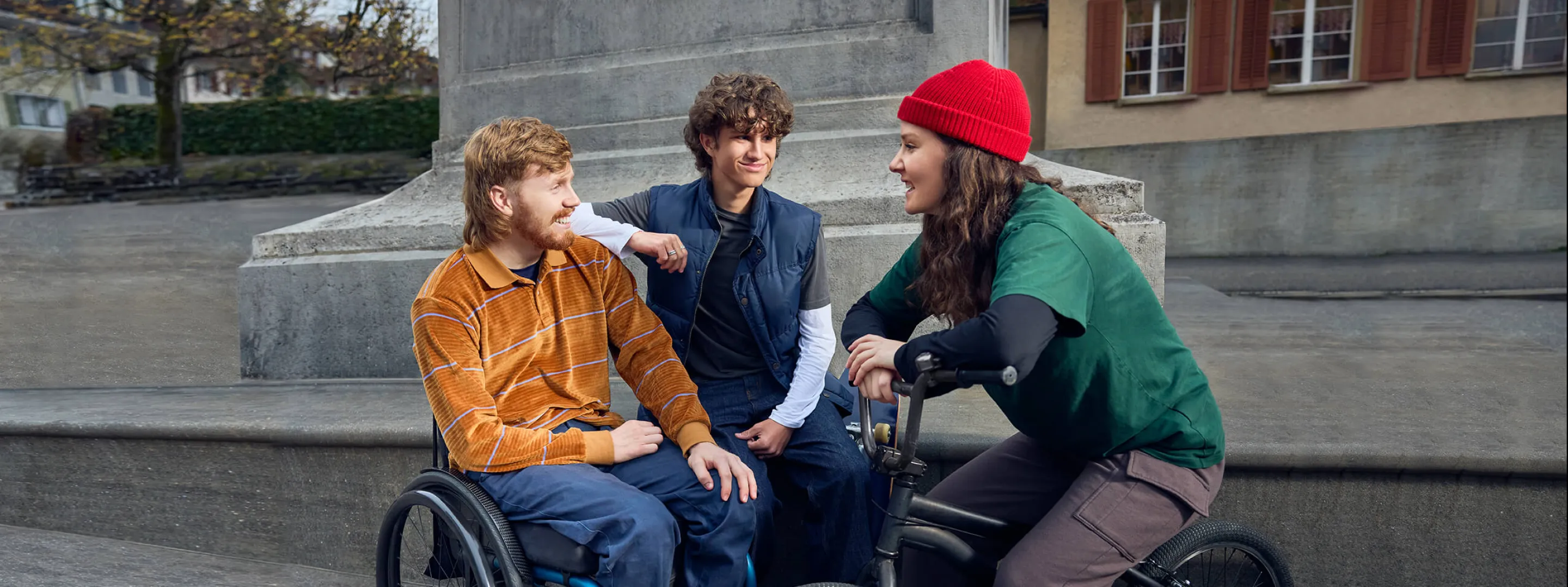 Tim Harder sitzt im Rollstuhl vor dem Sockel einer Statue zusammen mit zwei Freunden: Ein junger Mann sitzt auf dem Sockel und lehnt sich an Tims Schulter an, während eine junge Frau mit roter Mütze auf einem Mountainbike sitzt und zu den beiden jungen Männern blickt.