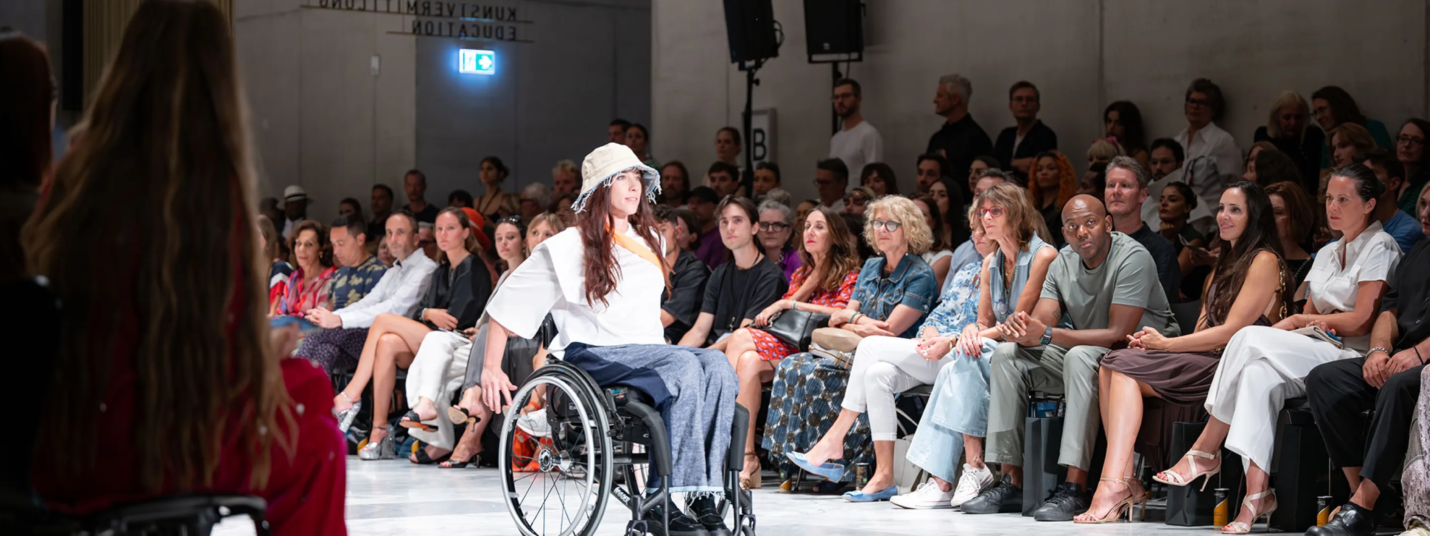 Modeshow mit Rollstuhlfahrerin auf dem Laufsteg die inklusive Mode trägt.