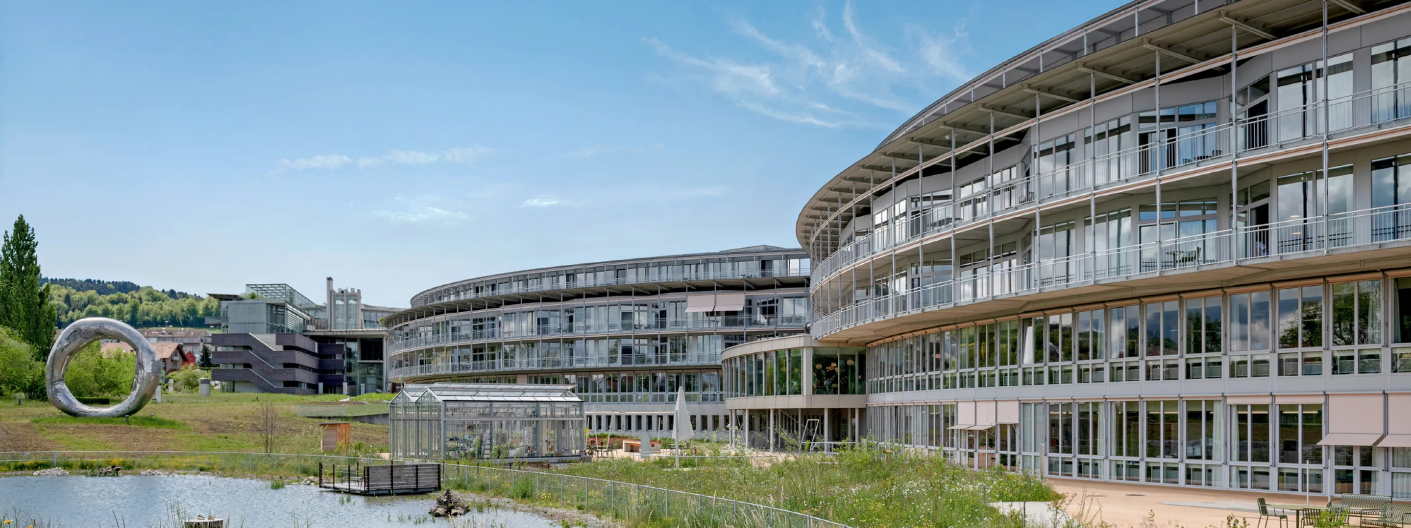 Das moderne Gebäude mit viel Glas und Holzvertäfelung, umgeben von einer Rasenfläche und einem kleinen See.