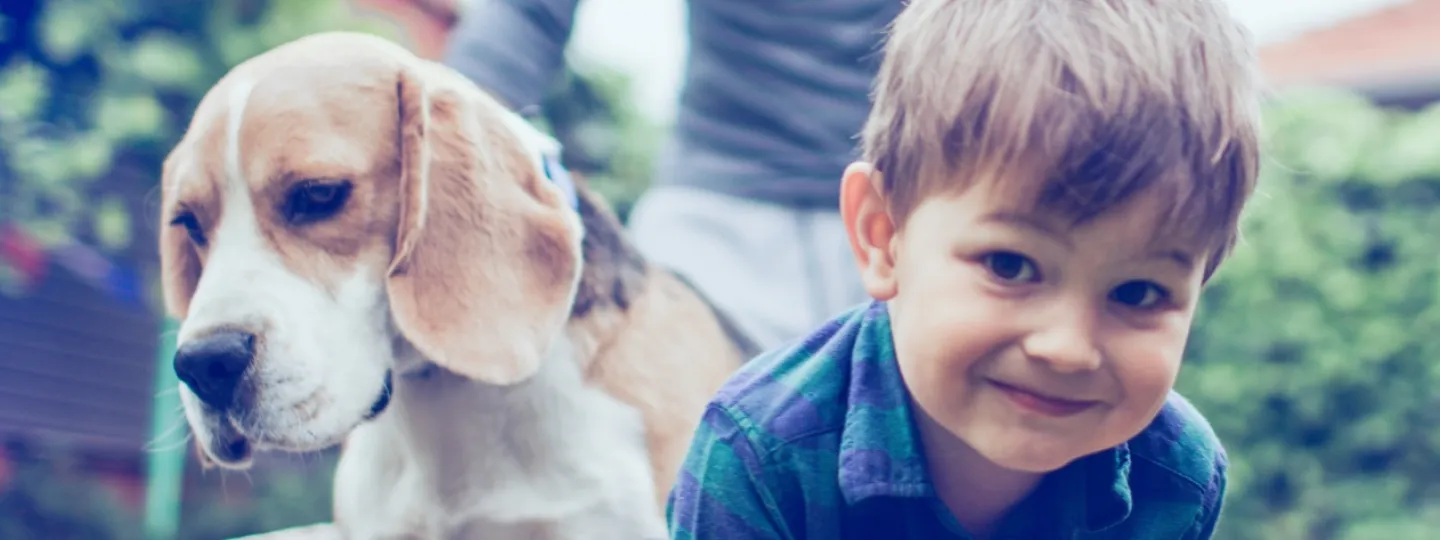 Un homme pousse une brouette dans un jardin. Dans la brouette se trouvent un enfant riant et un chien.  