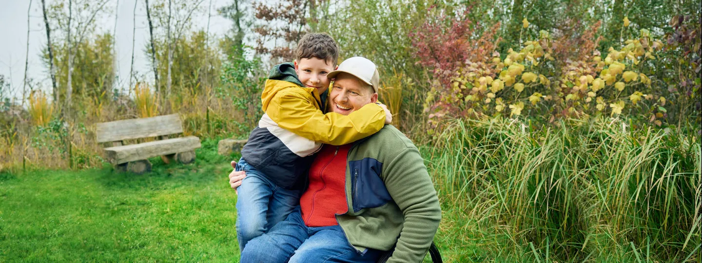 Heiri sitzt in seinem Rollstuhl auf einer Wiese und sein Sohn klettert ihm halbwegs auf den Schoss und umarmt seinen Vater ganz fest. Beide lächeln.
