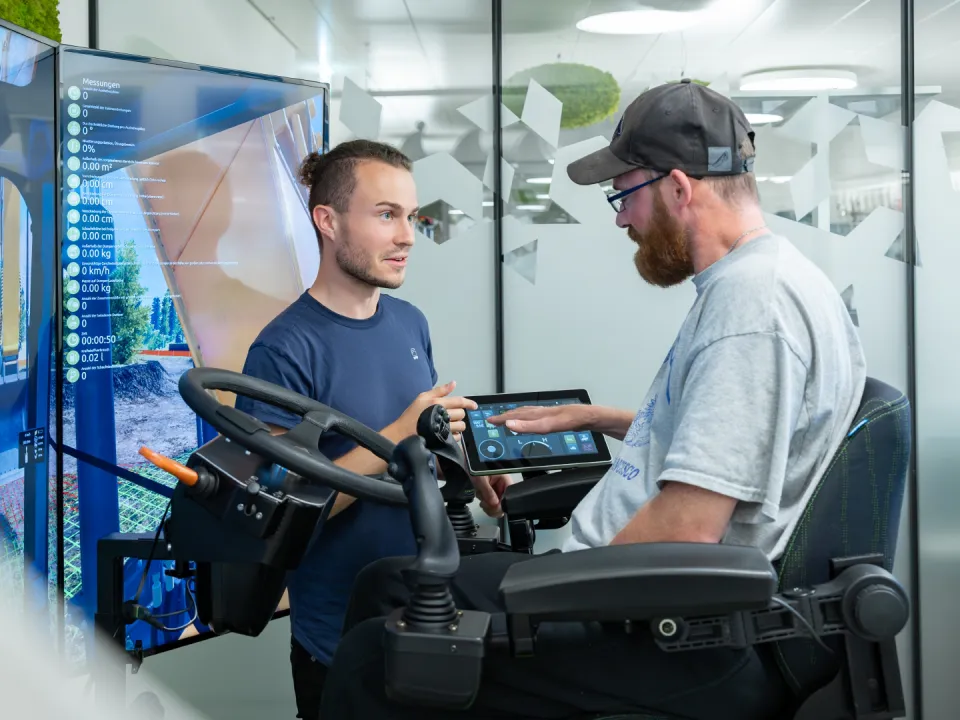 Ein Mann im Rollstuhl steuert einen Baumaschinensimulator und ein Mann erklärt es ihm.