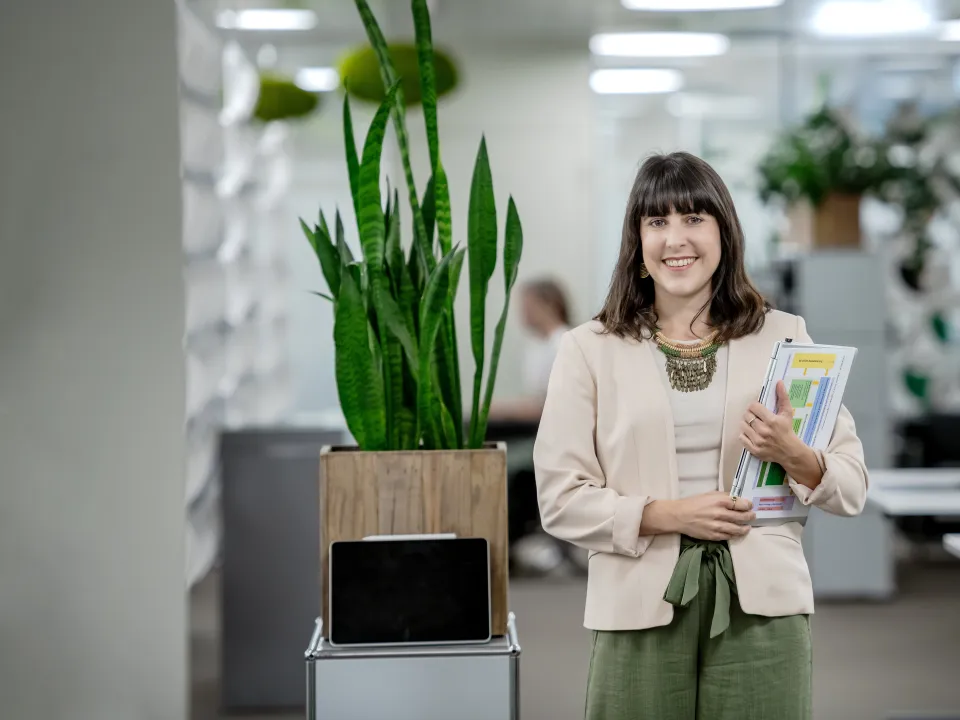 Selina Infanger steht lächelnd in einem modernen Büro. Sie trägt eine helle Jacke, ein beigefarbenes Oberteil und weite grüne Hosen. In ihren Armen hält sie mehrere Dokumente und Unterlagen. Im Hintergrund befinden sich Büropflanzen, darunter eine hohe Grünpflanze in einem Holztopf, der auf einem kleinen Regal steht, sowie weitere Dekorationselemente und Möbel. Die Büroeinrichtung wirkt hell und offen, mit modernen Gestaltungselementen und einer klaren, strukturierten Umgebung.