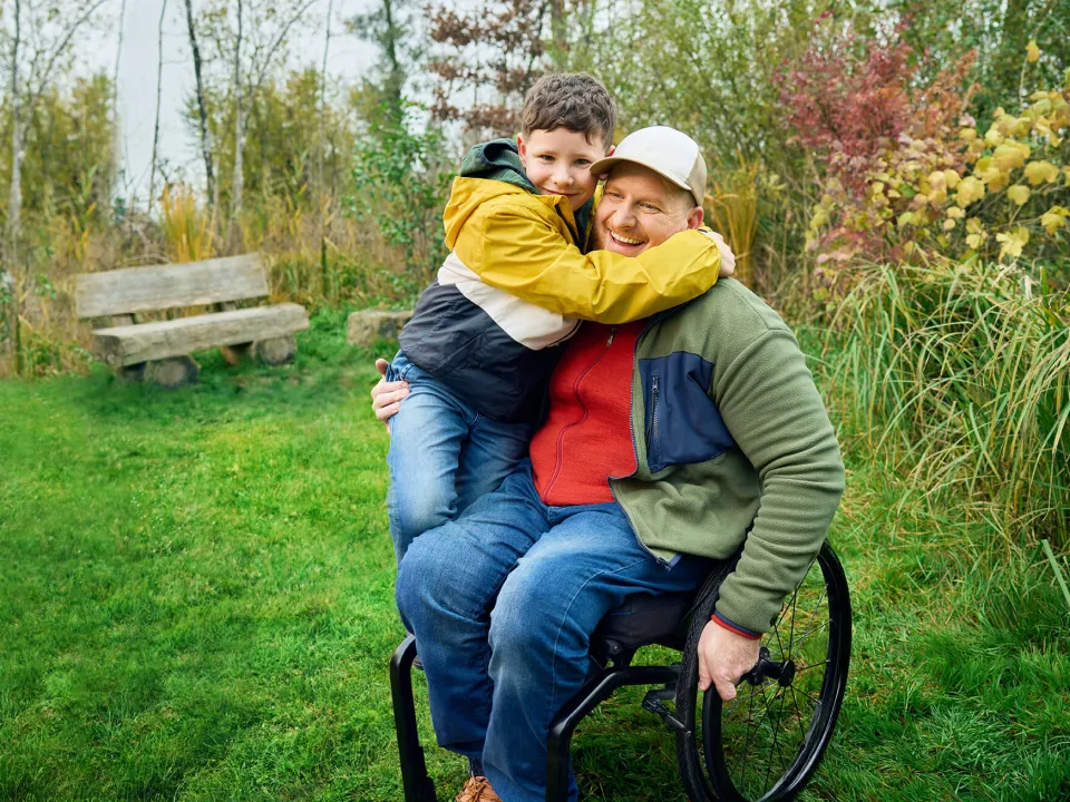 Heiri sitzt in seinem Rollstuhl auf einer Wiese und sein Sohn klettert ihm halbwegs auf den Schoss und umarmt seinen Vater ganz fest. Beide lächeln.