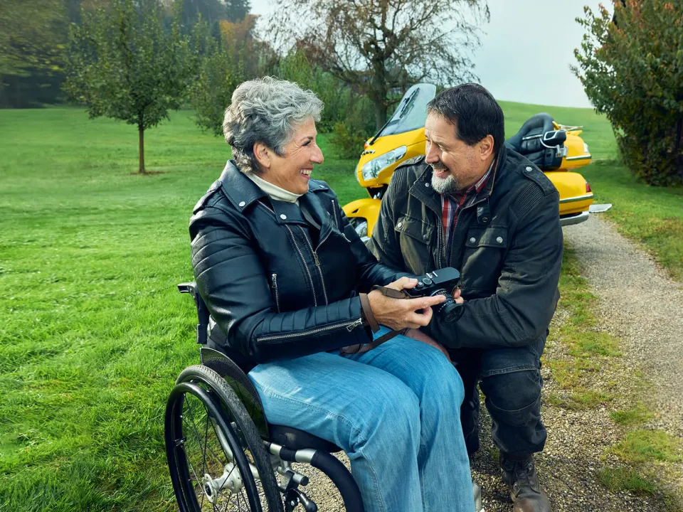 Rosa Zaugg sitzt im Rollstuhl auf einem Kiesweg, hält eine Kamera in der Hand und lächelt ihren Partner Lukas an.