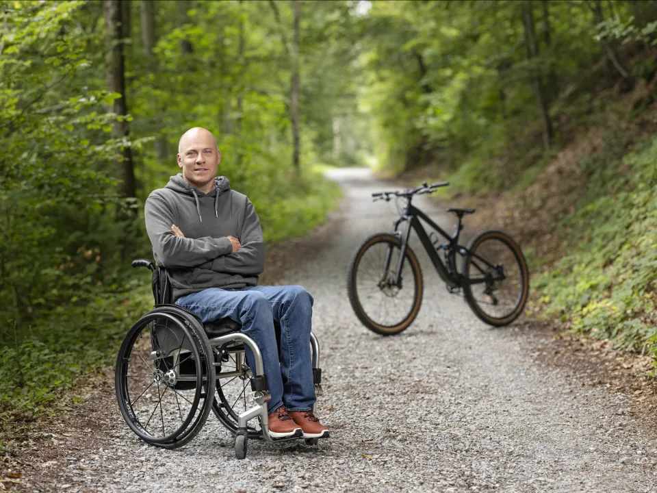 Sebastian Tobler sitzt in seinem Rollstuhl im Wald und lächelt mit verschränkten Armen in die Kamera. Im Hintergrund ist ein Mountain Bike zu sehen. 