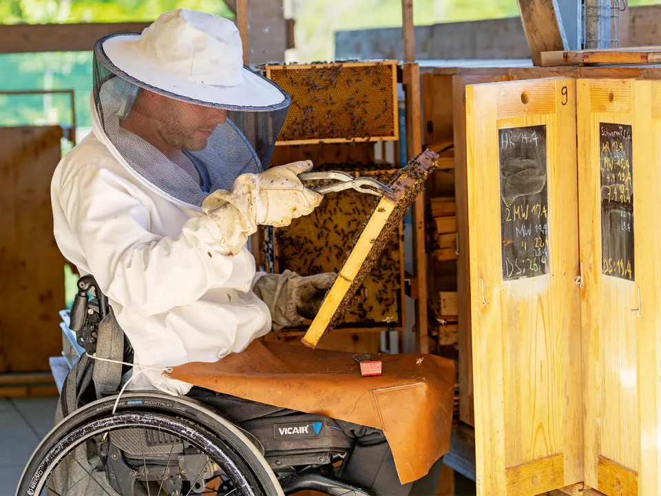 Markus Buser sitzt im Rollstuhl und trägt einen Bienenschutz. In der Hand hat er eine Wabe.