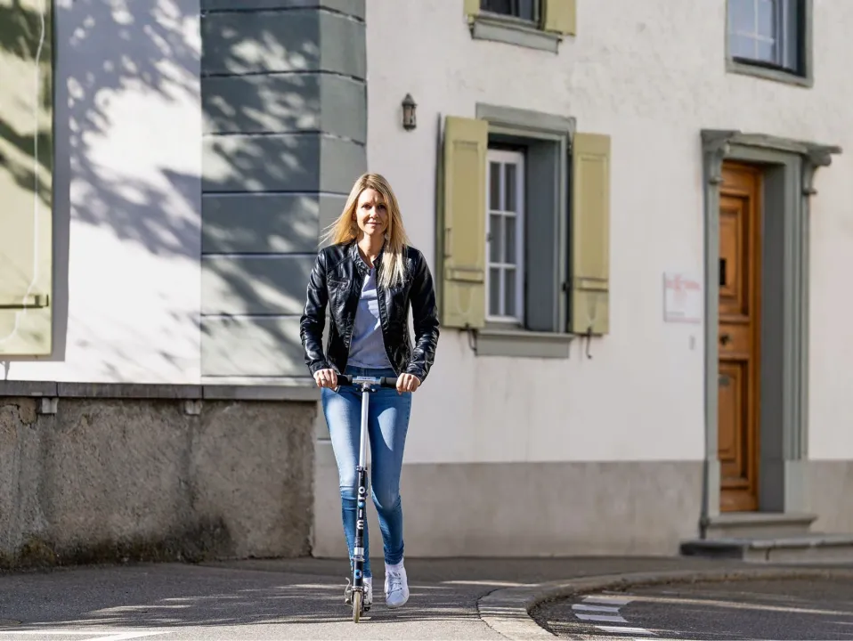 Eine Frau fährt mit einem Trottinett durch eine ruhige Stadtstrasse vor einem historischen Gebäude – selbstständige Mobilität, urbane Barrierefreiheit und aktive Teilhabe im Alltag.