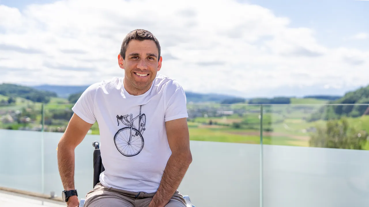 A young man in a white t-shirt smiling while sitting on a balcony with a scenic mountain landscape in the background.