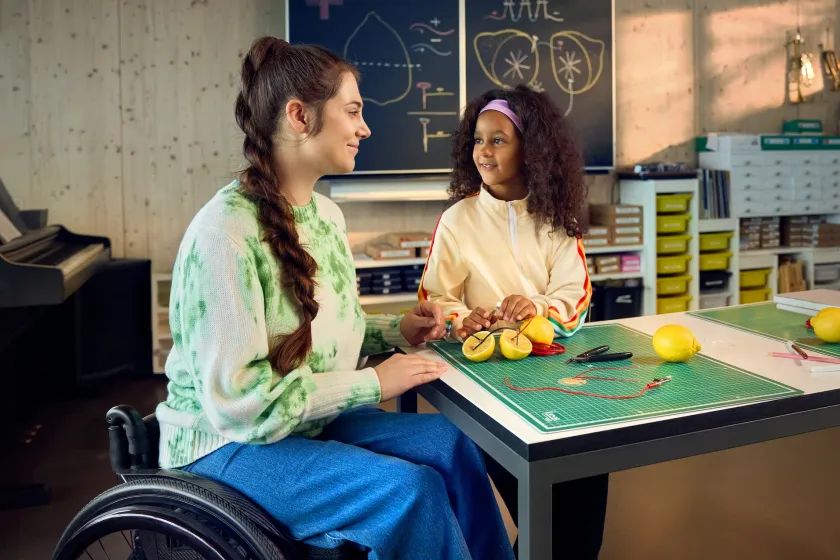 Chiara Schlatter sitzt im Rollstuhl an einem Tisch in einem Schulzimmer zusammen mit einer Schülerin und zeigt ihr, wie man mit Zitronen eine Lampe zum Leuchten bringen kann. Beide schauen sich an und lächeln.
