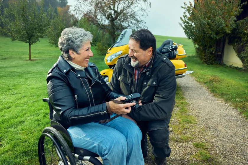 Rosa Zaugg sitzt in ihrem Rollstuhl auf einem Feldweg auf einem Bauernhof und blickt zu ihrem Partner Lukas hinüber, der sich neben ihr hingekniet hat. Sie zeigt ihm eine Kamera. Im Hintergrund ist seine gelbe Goldwing zu sehen.
