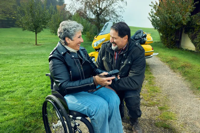 Rosa Zaugg sitzt im Rollstuhl auf einem Kiesweg, hält eine Kamera in der Hand und lächelt ihren Partner Lukas an.