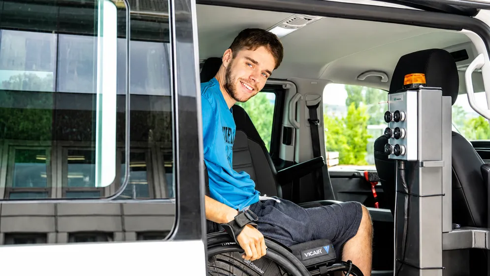 Smiling man in blue shirt seated inside vehicle, holding steering wheel.