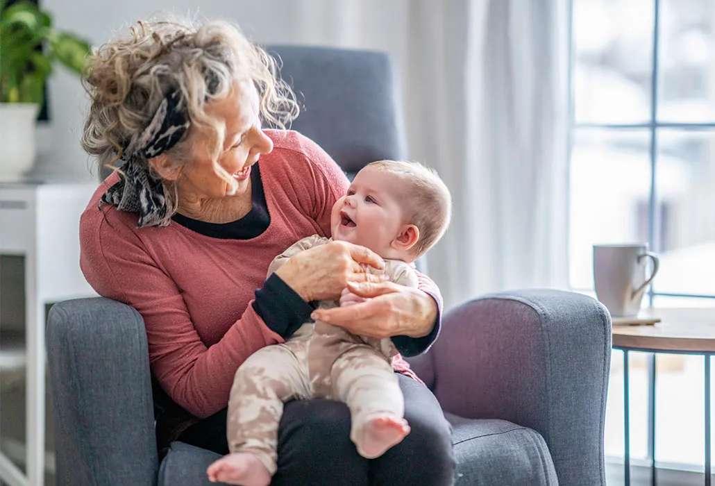 An older woman is holding a smiling baby in her arms. The woman is sitting in an armchair.