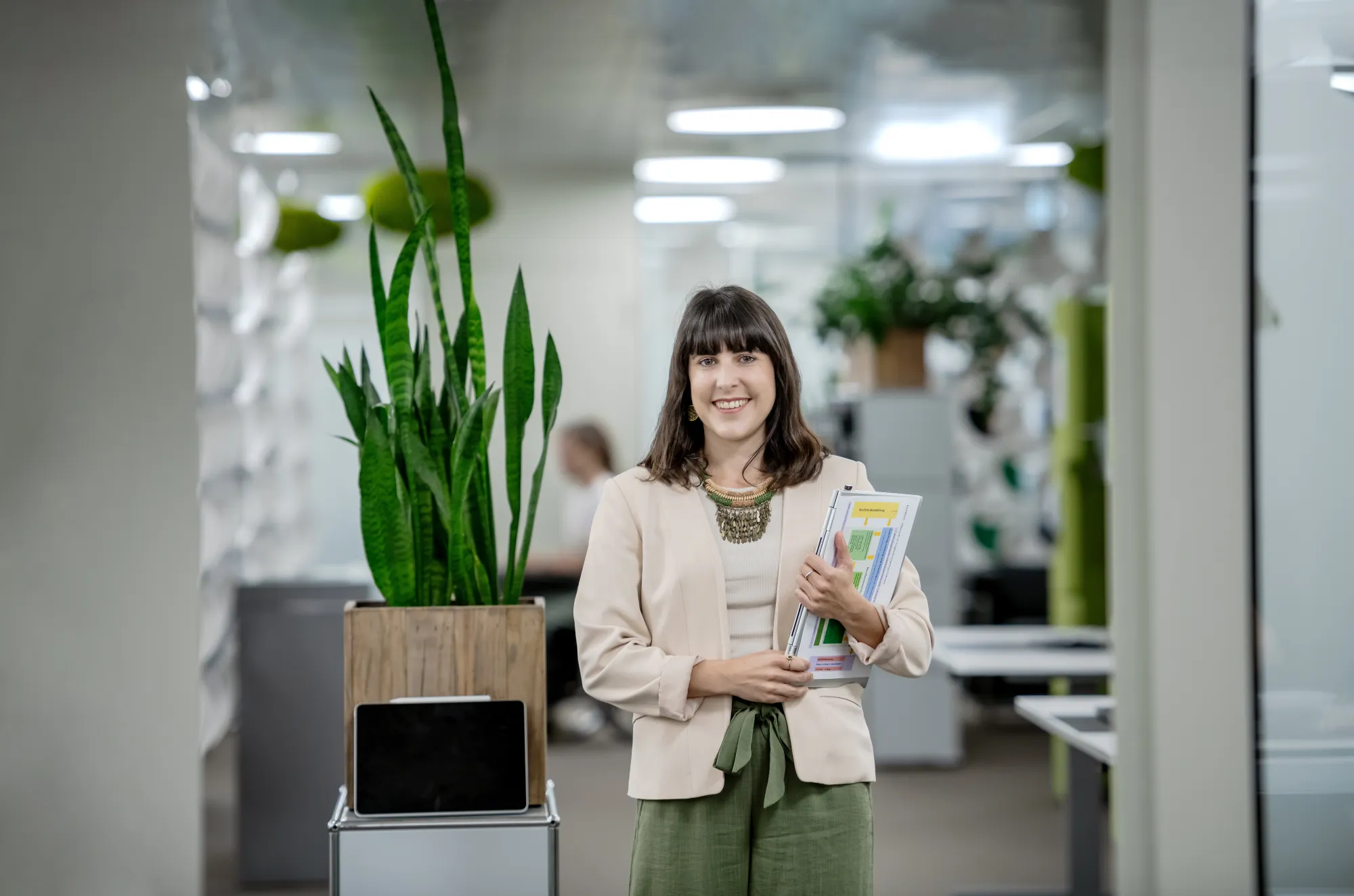Selina Infanger steht lächelnd in einem modernen Büro. Sie trägt eine helle Jacke, ein beigefarbenes Oberteil und weite grüne Hosen. In ihren Armen hält sie mehrere Dokumente und Unterlagen. Im Hintergrund befinden sich Büropflanzen, darunter eine hohe Grünpflanze in einem Holztopf, der auf einem kleinen Regal steht, sowie weitere Dekorationselemente und Möbel. Die Büroeinrichtung wirkt hell und offen, mit modernen Gestaltungselementen und einer klaren, strukturierten Umgebung.