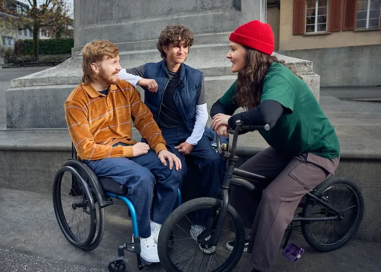 Tim Harder sitzt im Rollstuhl vor dem Sockel einer Statue zusammen mit zwei Freunden: Ein junger Mann sitzt auf dem Sockel und lehnt sich an Tims Schulter an, während eine junge Frau mit roter Mütze auf einem Mountainbike sitzt und zu den beiden jungen Männern blickt.