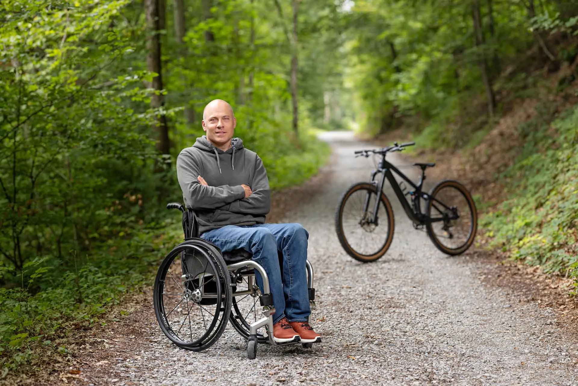Sebastian Tobler est en fauteuil roulant dans la forêt et sourit dans la caméra, les bras croisés. À l’arrière-plan se trouve un VTT. 