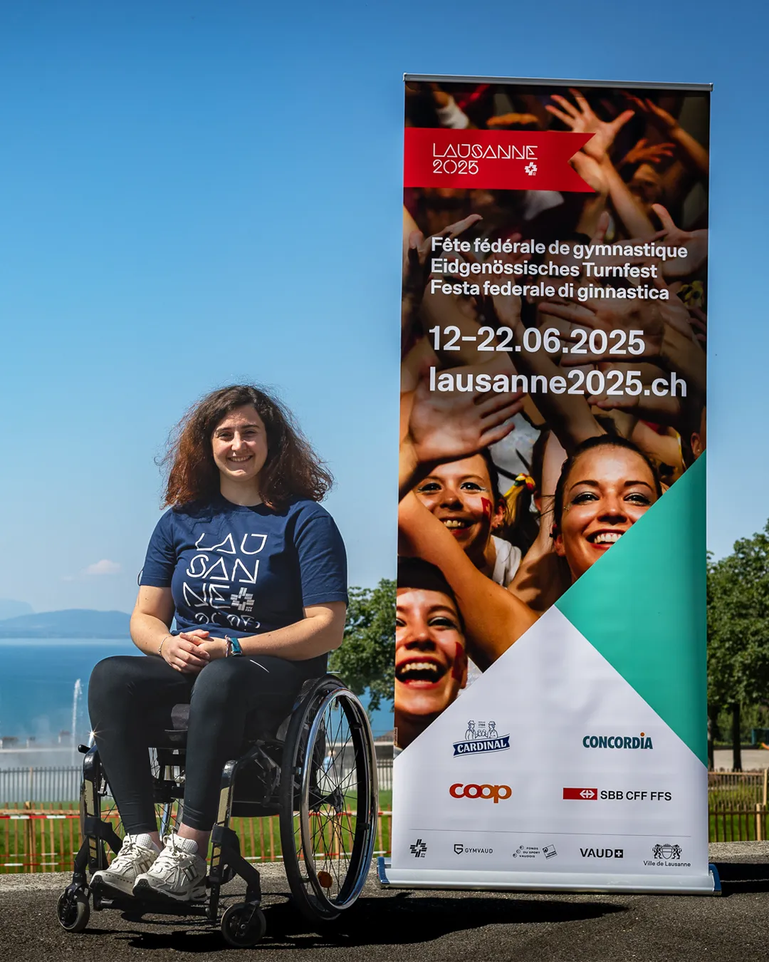 Giulia Damiano sitzt in ihrem Rollstuhl vor dem Genfersee und trägt ein T-Shirt mit der Aufschrift "Lausanne 2025". Rechts von ihr ist die Werbung für das Eidgenössische Turnfest in Lausanne. 