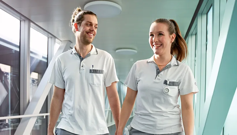 Zwei Pflegefachkräfte gehen zufrieden einen Spitalgang entlang. Paraplegie Juni 2025 Teaser
