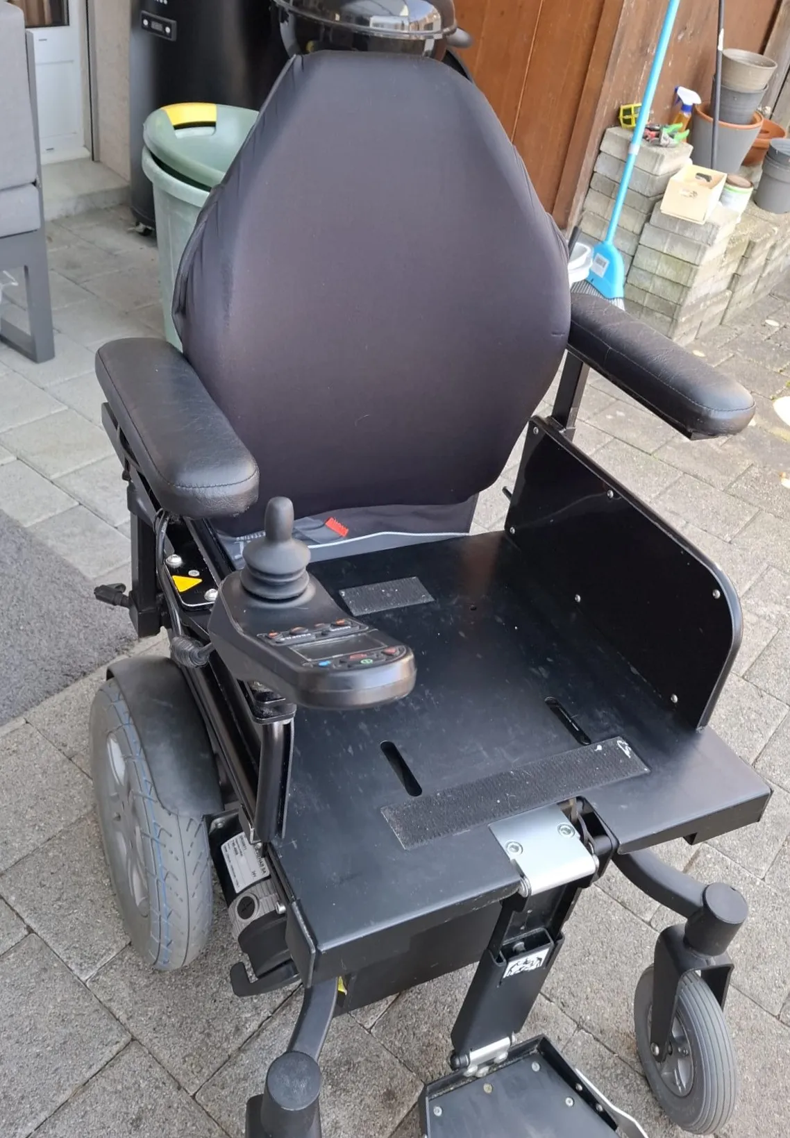 Elektrischer Rollstuhl mit grauem Sitz und Armlehnen, auf Holzboden stehend.