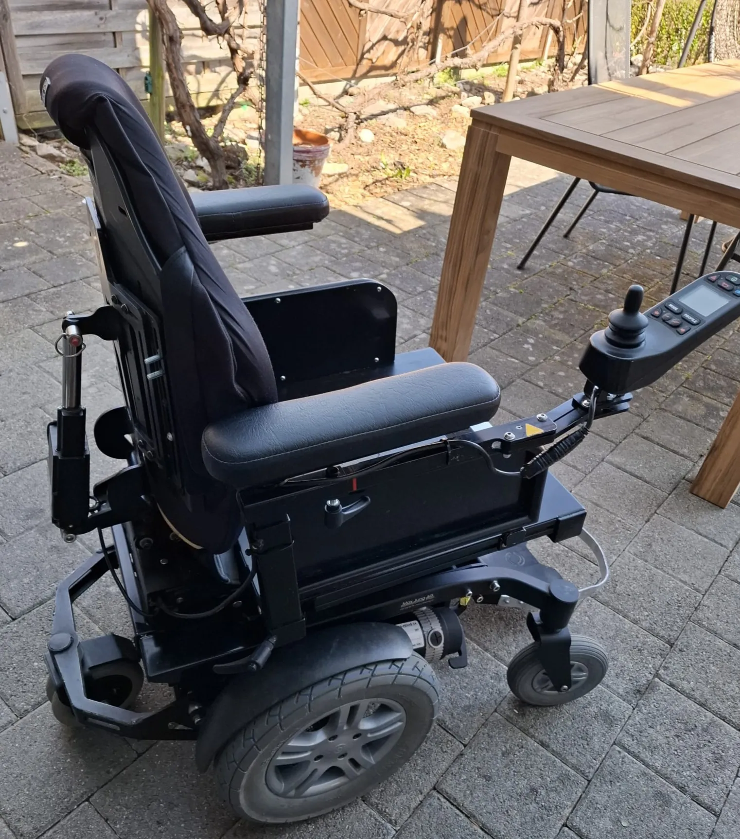 Elektrischer Rollstuhl mit schwarzem Sitz auf Holzterrasse neben Tisch.