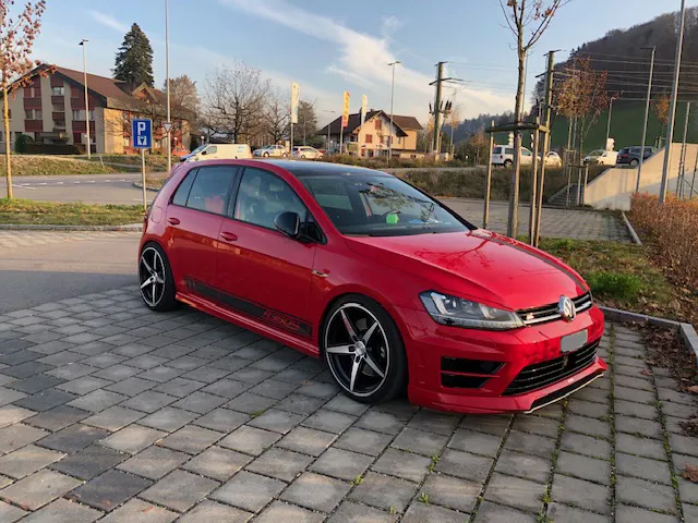 Roter Volkswagen Golf GTI parkt auf gepflasterter Straße vor Häusern und Bäumen.