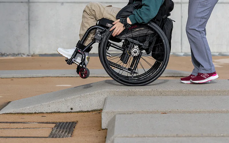 Rollstuhl fährt eine Rampe hinunter, im Vordergrund ist eine Treppe