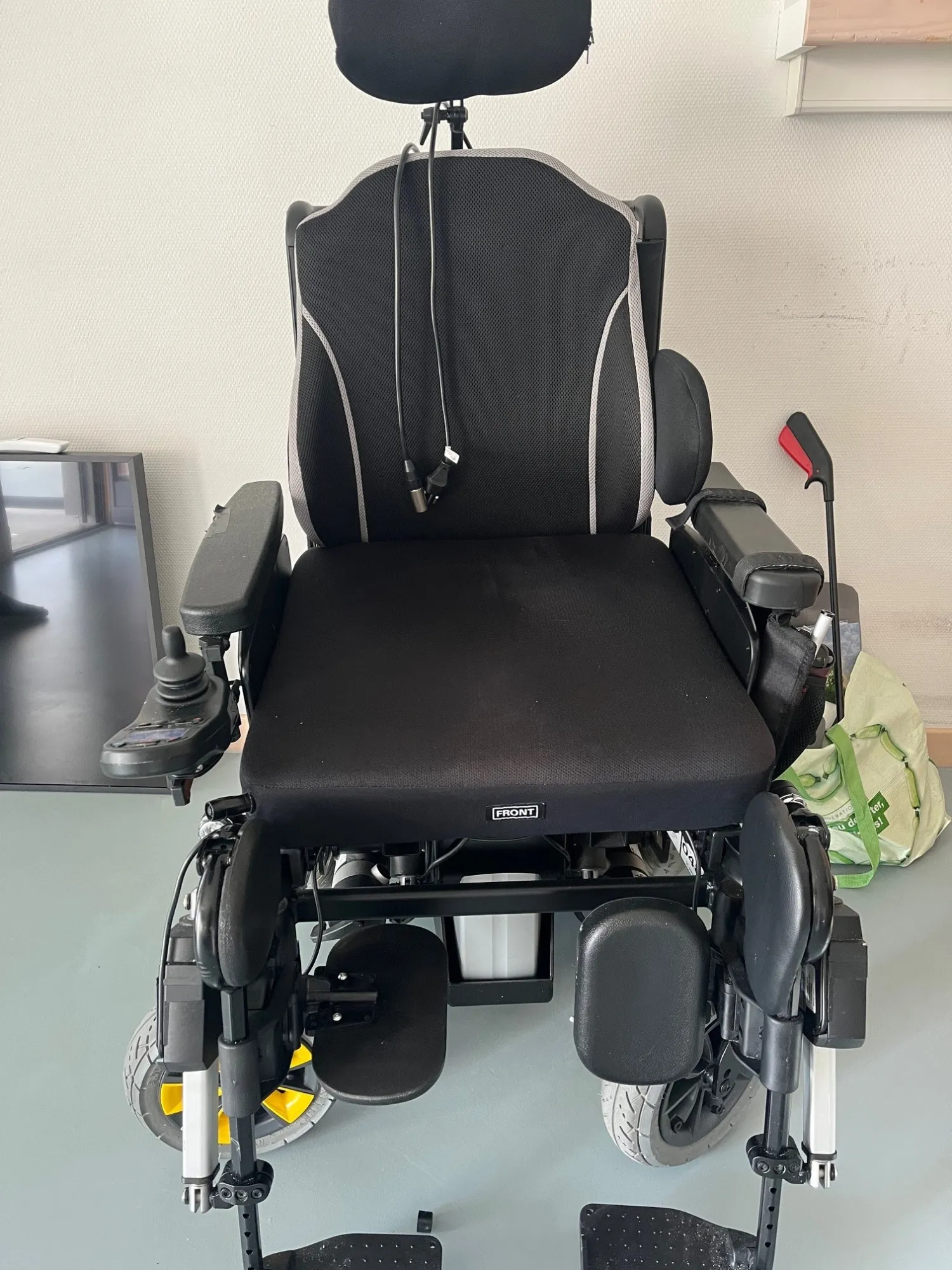 Schwarzer elektrischer Rollstuhl mit Kopfstütze und Armlehnen vor heller Wand.