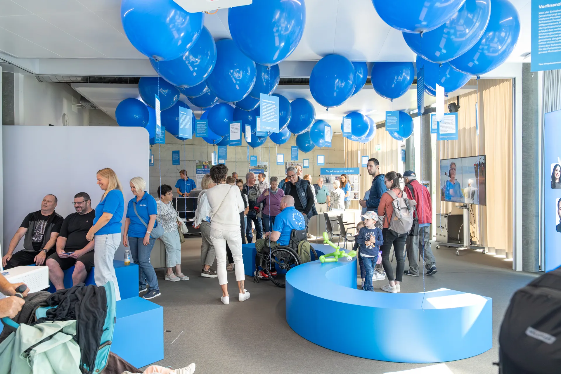 Stand der Schweizer Paraplegiker-Stiftung an den Tagen der offenen Tür der Schweizer Paraplegiker-Gruppe, mit vielen Besucherinnen und blauen Ballonen, die an der Decke hängen. 