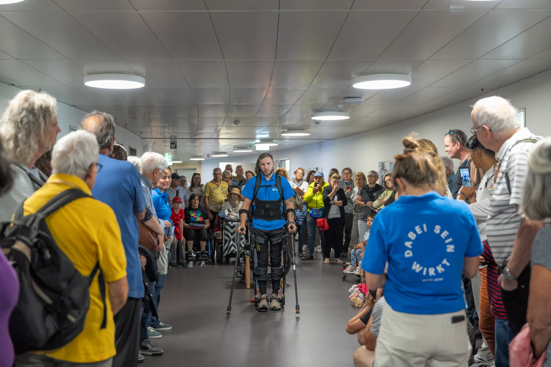 Besucherinnen der Tage der offenen Tür in Nottwil beobachten, wie ein Mann mit Hilfe eines Exoskeletts an Krücken geht.